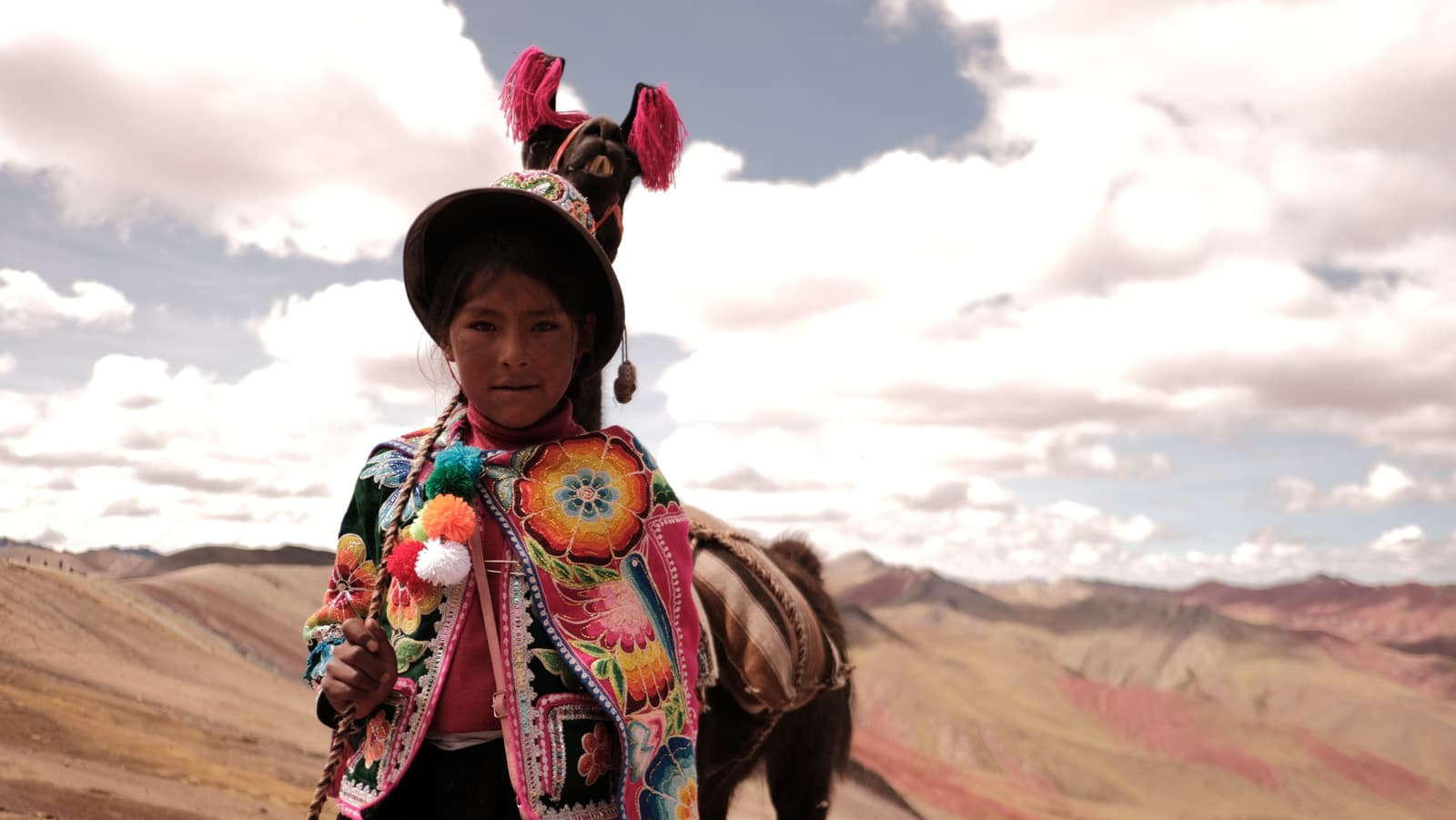 Young girl in vibrant embroidered clothing with llama, rainbow mountains behind